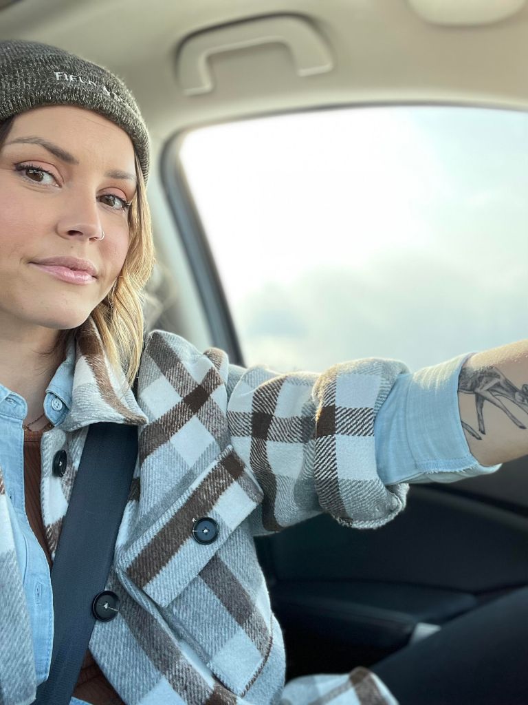 A photo of the blogger megdanielle in her car with her hand draped over the steering wheel. She wears a beanie and a plaid jacket, her tattoo peaking past rolled sleeves.